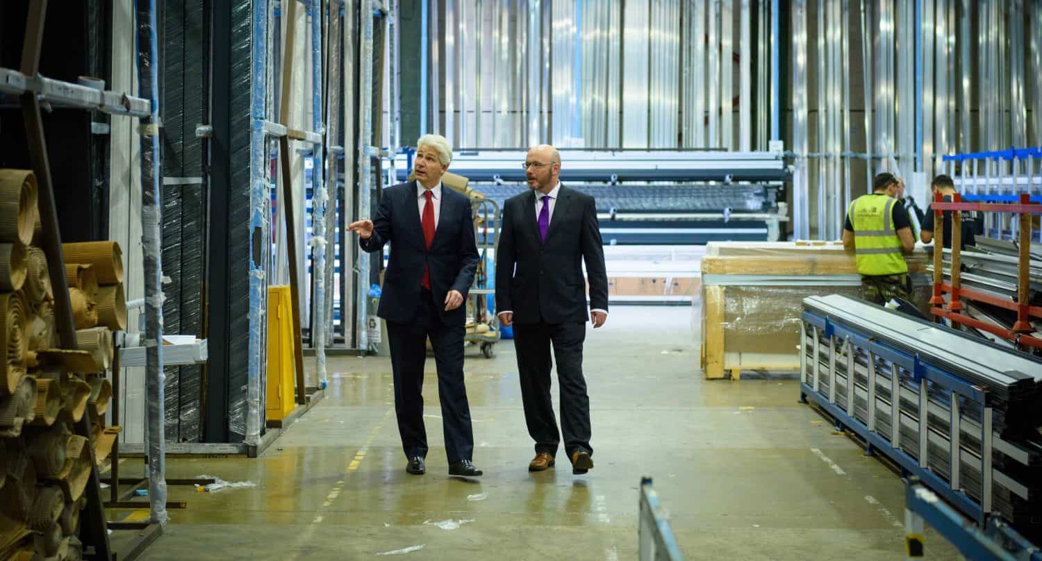 Two men in suits walk and talk through a spacious warehouse with metal beams and wooden materials, discussing bespoke glass solutions, while a worker in a yellow safety vest organizes shelves in the background.