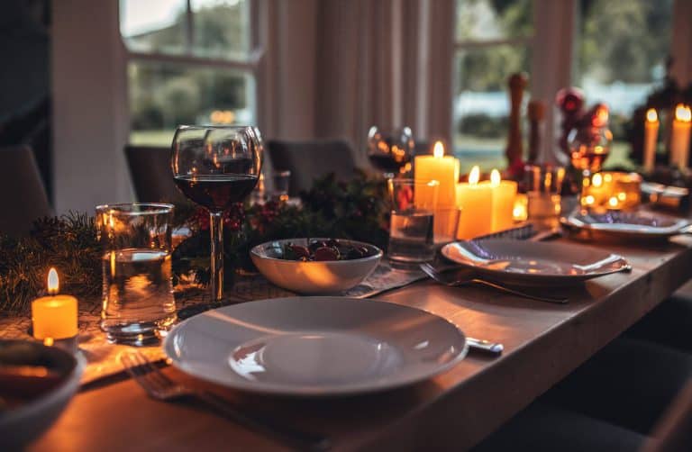 A warmly lit dining table set for a meal with candles, plates, cutlery, wine glasses, water glasses, and a bowl of cherries, with a festive garland as decoration in a cozy room with large windows.