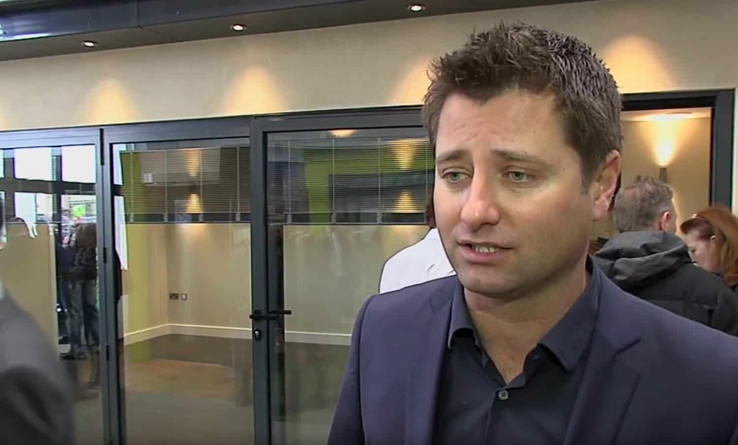 A man in a dark suit stands indoors, speaking with a serious expression. Glass doors and several people are visible in the background, suggesting a modern office or event setting.