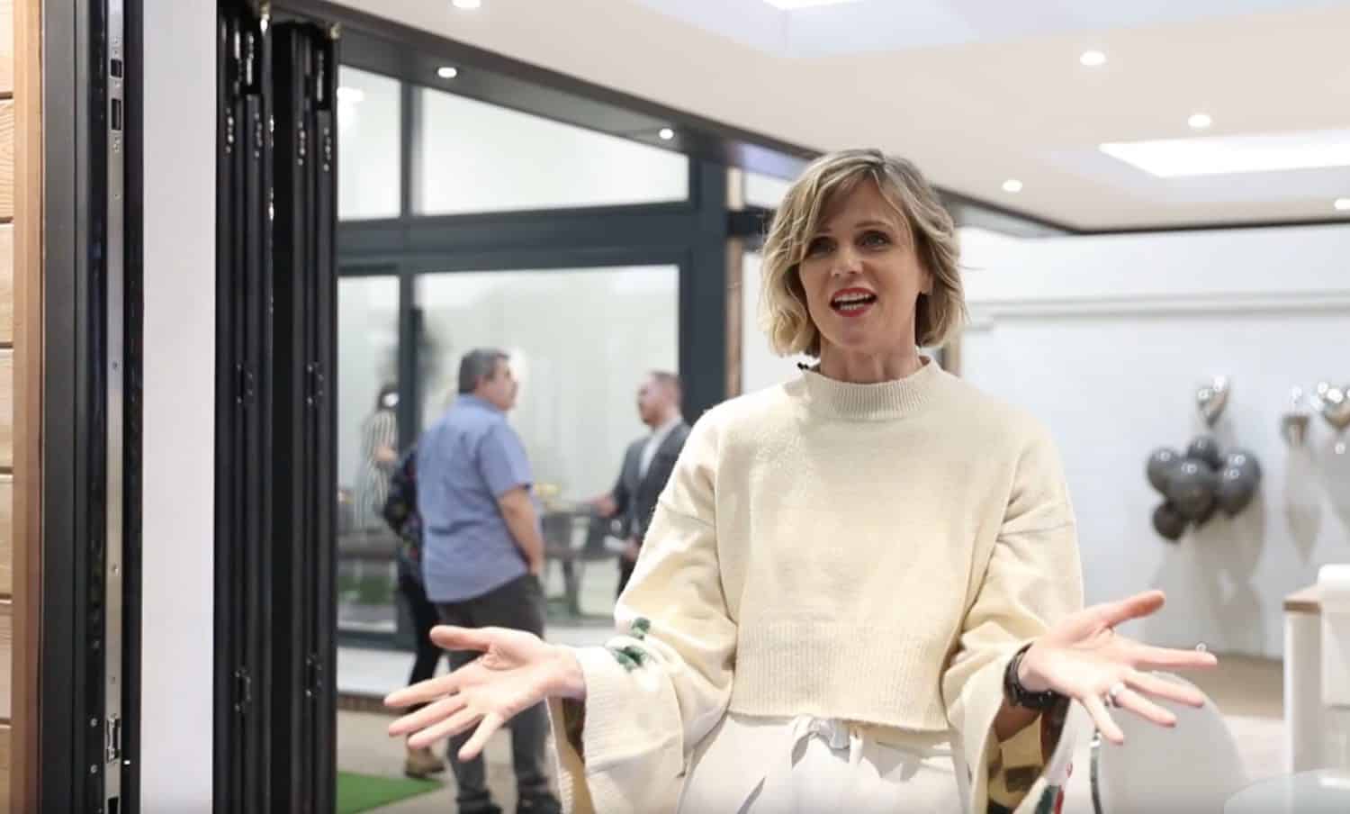 A woman with short blonde hair, wearing a light sweater, stands indoors with her arms open, speaking or presenting. In the background, there are several people, large windows, and silver balloons.