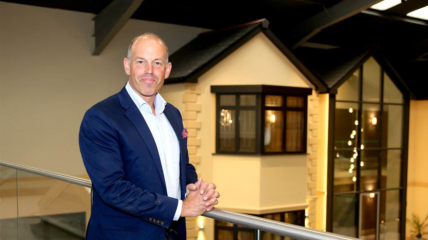 A man in a navy blue suit and white shirt stands indoors by a glass railing, smiling, with a modern house-like structure and large windows in the background.