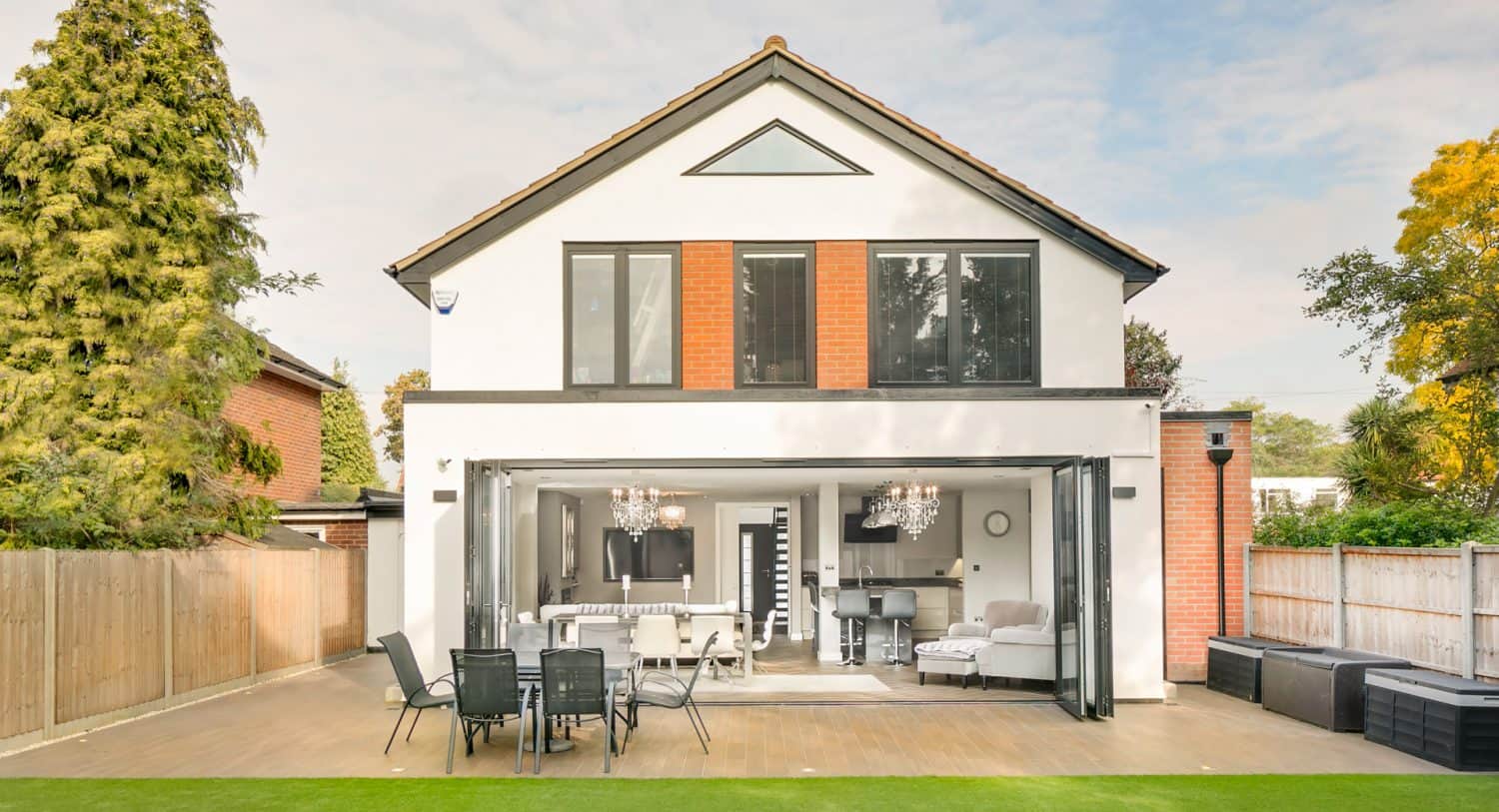 A modern two-story house with large windows and folding glass doors opening to a patio with outdoor dining furniture, surrounded by trees and a fenced yard.
