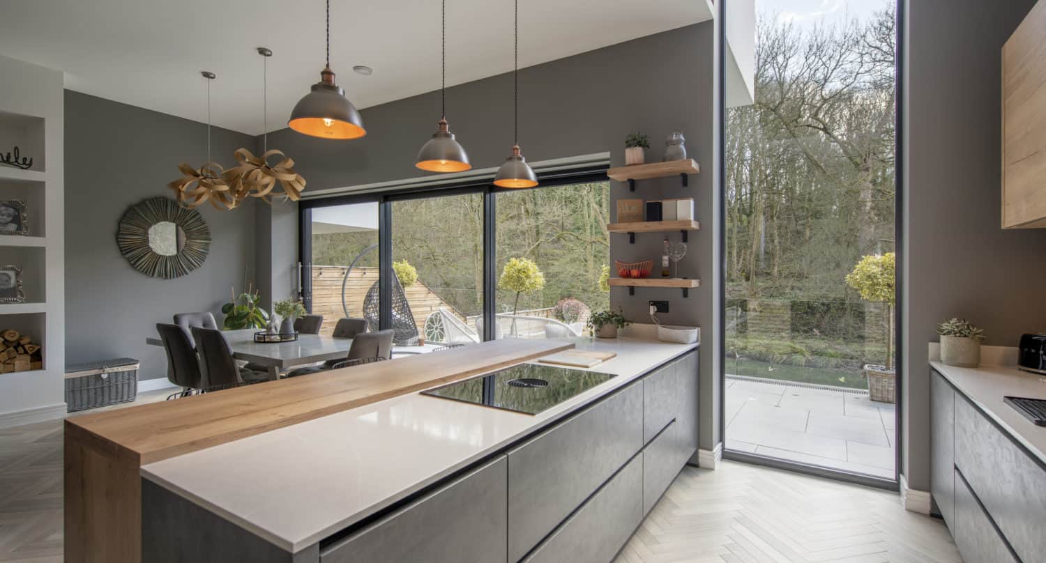 Modern kitchen with gray cabinets, a wooden countertop island, pendant lights, and large floor-to-ceiling bifold doors overlooking the patio and trees. Dining area with a round table is visible in the background.