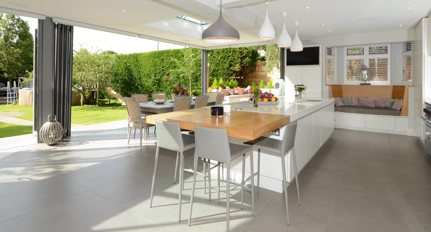 Modern open-plan kitchen with a large island, dining table, and seating area. Glass doors open to a green garden with outdoor seating, creating a bright and spacious indoor-outdoor living space.