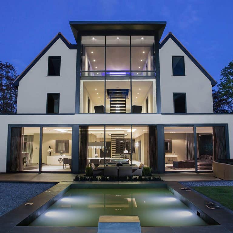 Modern three-story house with large glass windows, open-plan design, and interior lights on at dusk. A sleek outdoor pool is in the foreground, reflecting the buildings contemporary architecture.
