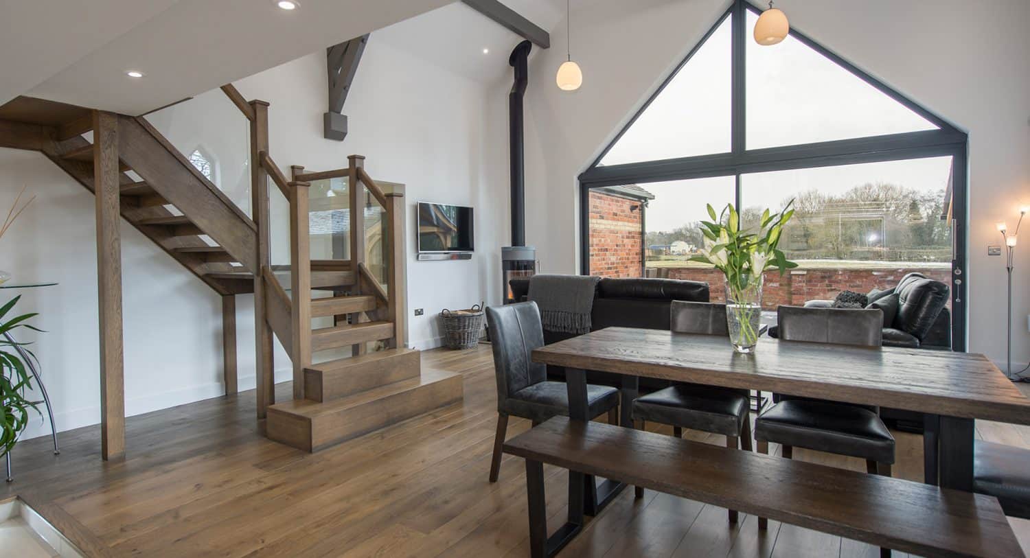 Modern open-plan living and dining area with large triangular window, wooden staircase, dark wood furniture, black chairs and bench, and a vase of flowers on the table. Natural light fills the room.
