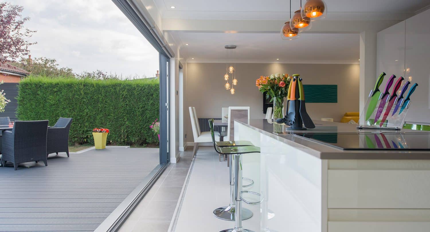 Modern open-plan kitchen with glossy white cabinets and a breakfast bar opens onto a patio with outdoor dining, separated by large sliding glass doors. Pendant lights and colorful knives add stylish accents.