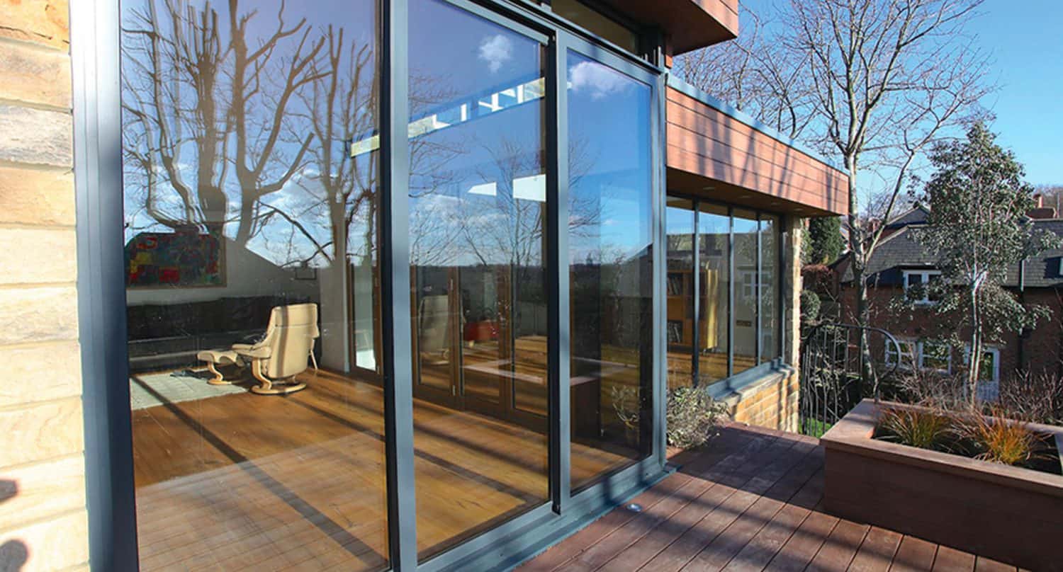 Large glass sliding doors reflect leafless trees and blue sky, leading from a wooden deck into a modern interior with wooden floors and a cream chair. A planter and nearby houses are visible outside.
