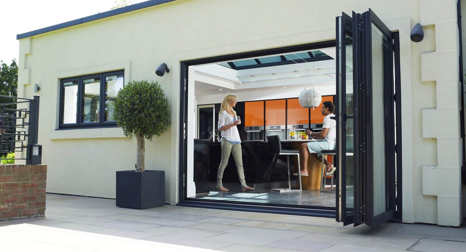 A modern home with large glass folding doors open to a patio. Inside, a woman stands while another person sits at a kitchen island, talking. There is a potted tree outside near the doorway.