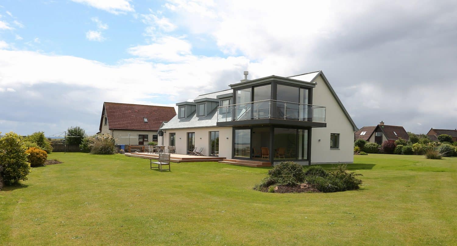 Modern two-story house with large windows and a glass balcony, surrounded by a spacious, well-maintained green lawn and some shrubs, under a partly cloudy sky.