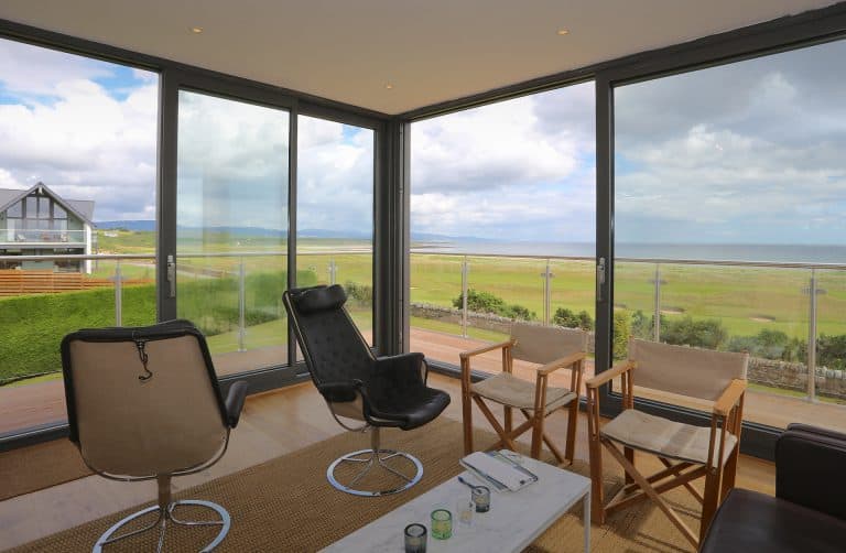 Modern sitting area with black and tan chairs facing a coffee table, surrounded by large glass windows offering expansive views of grassy fields, a distant house, and the ocean under a cloudy sky.