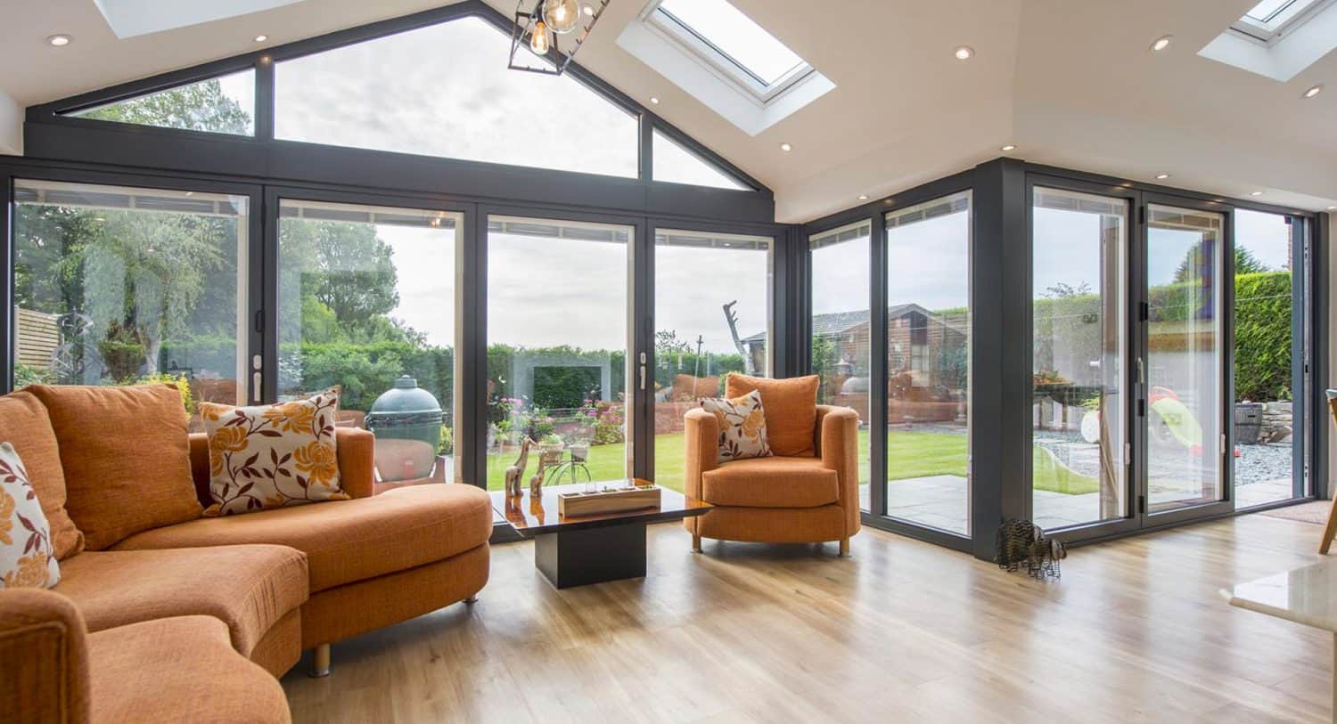 A bright, modern sunroom with large floor-to-ceiling glass doors, orange sofas with patterned cushions, a wooden coffee table, and views of a green garden outside.