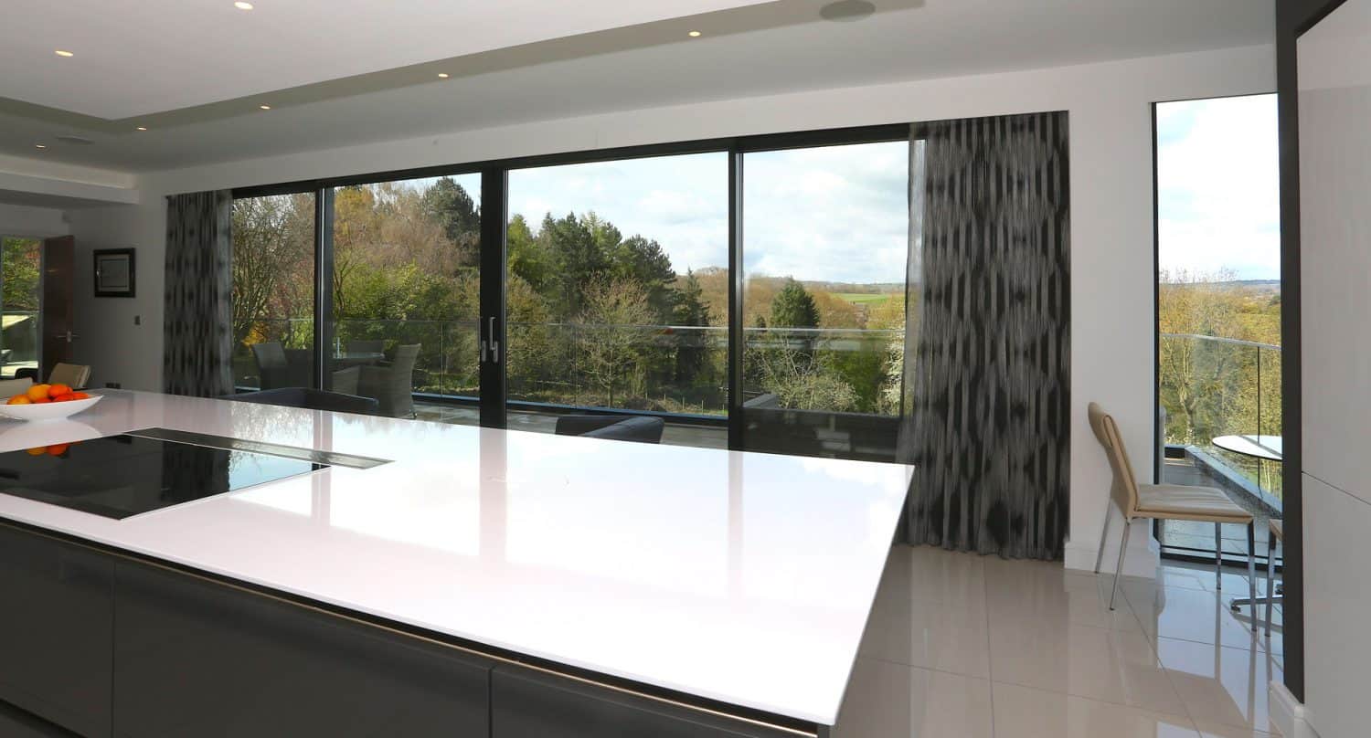 Modern kitchen with a glossy white island countertop, built-in stovetop, and a bowl of fruit. Large sliding glass doors open to a balcony with a scenic view of trees and distant hills. Two chairs are placed by the window.