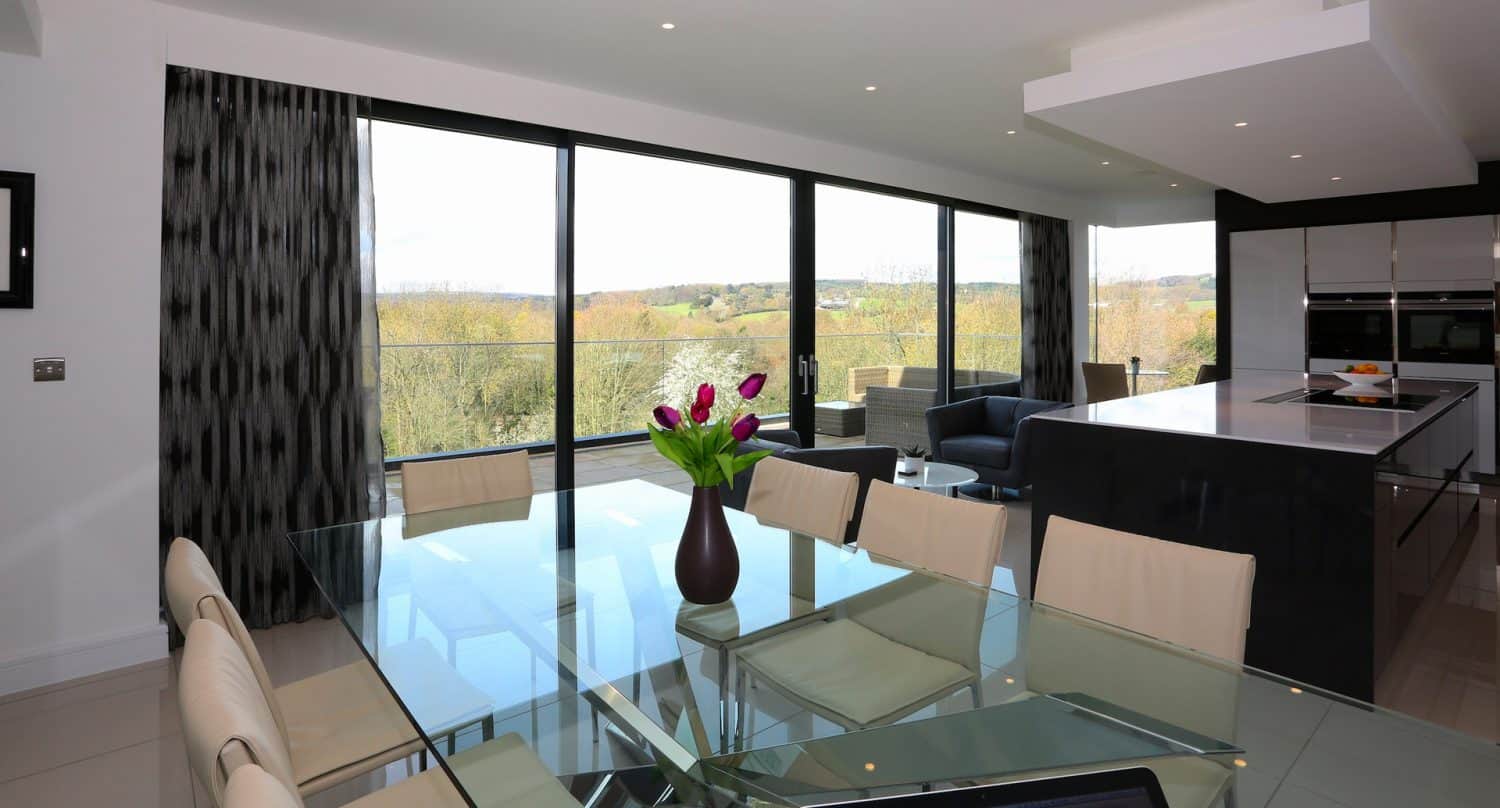 Modern dining and kitchen area with a glass table, cream chairs, a vase of tulips, and large floor-to-ceiling windows offering a scenic view of trees and a balcony.