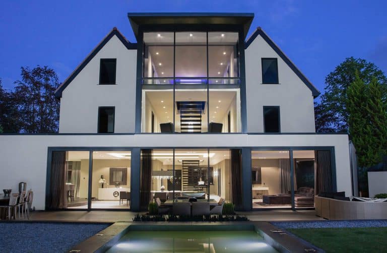 Modern three-story house with large glass windows, illuminated interiors, and a central staircase. The backyard features a small pool, outdoor seating, and landscaped greenery, all set at dusk.