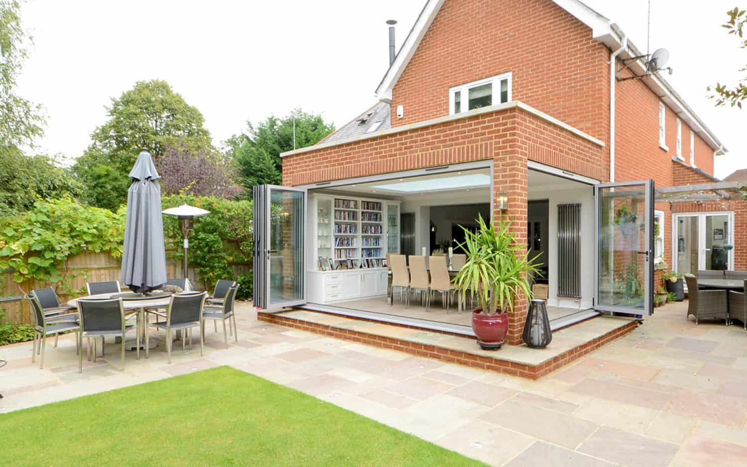 A modern brick house with large glass doors open to a patio. There’s a dining area inside, a table with chairs and umbrella outside, potted plants, and a well-kept lawn surrounded by greenery.