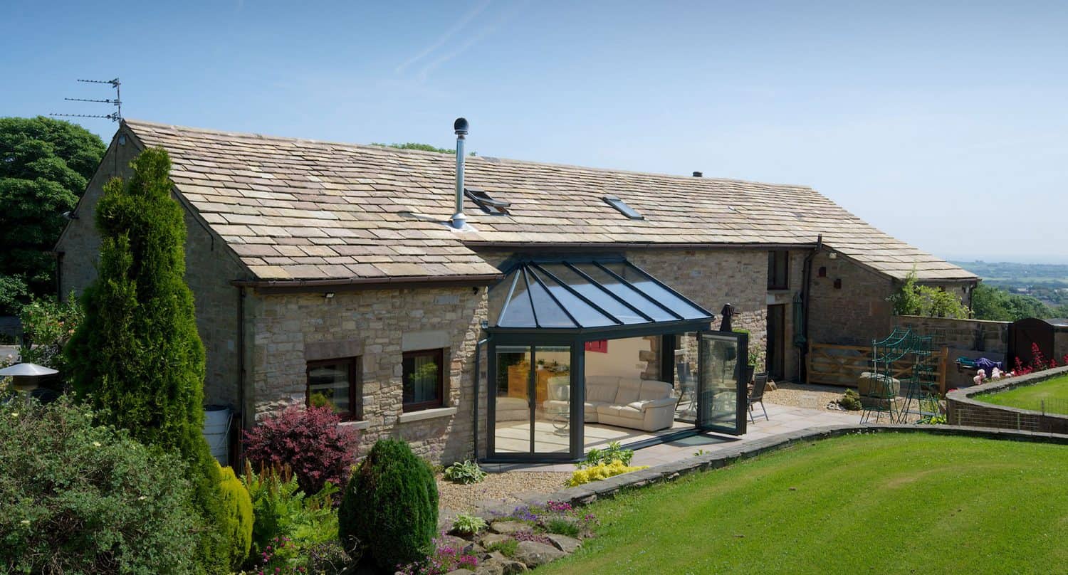 A large stone house with a glass sunroom extension featuring bespoke glass solutions, surrounded by a well-kept lawn, shrubs, and colorful garden beds, set against a clear blue sky and distant countryside views.