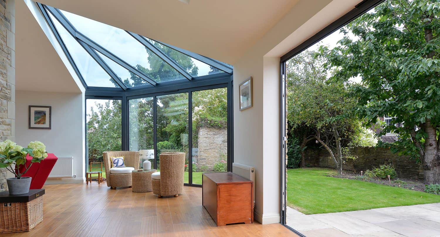 A modern sunroom with large glass windows and a glass ceiling opens onto a green garden. There are wicker chairs and a small table on the wooden floor, letting in lots of natural light.