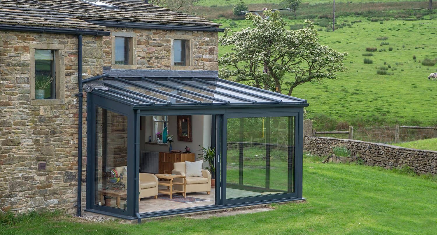 Modern glass conservatory attached to a stone house, featuring large windows, a glass roof, and light-colored furniture inside. The structure overlooks a green lawn and a rural landscape with trees and rolling hills.