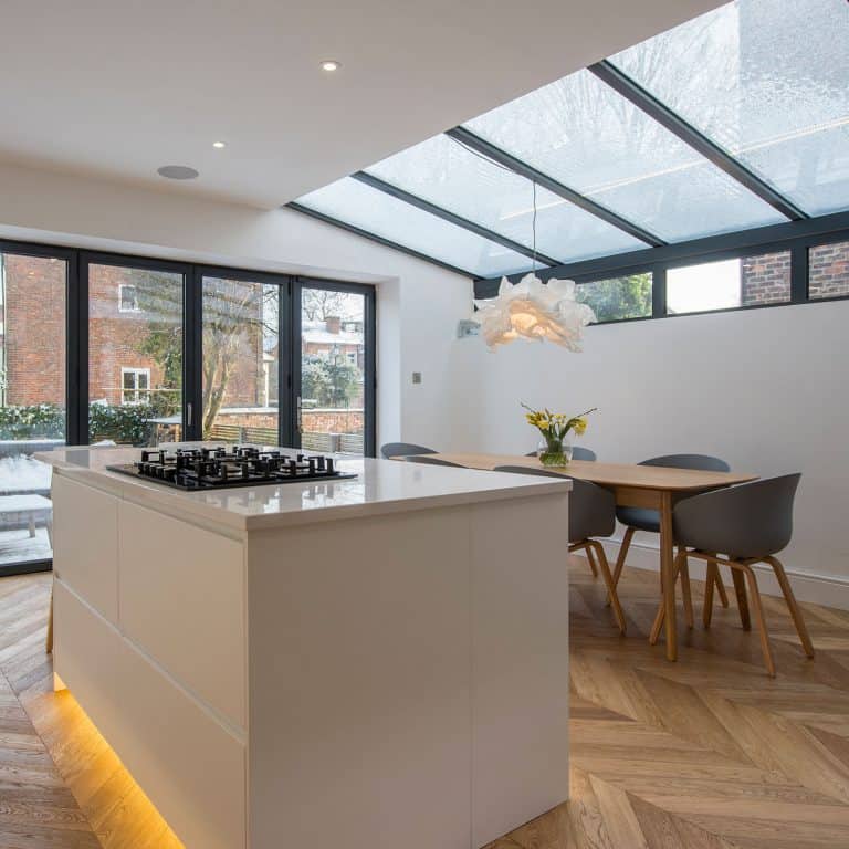 Modern kitchen with a white island and built-in gas stove, wooden dining table with gray chairs, herringbone floor, and bifold doors opening to the garden. A skylight ceiling and bespoke glass solutions fill the space with natural light. Yellow flowers adorn the table.