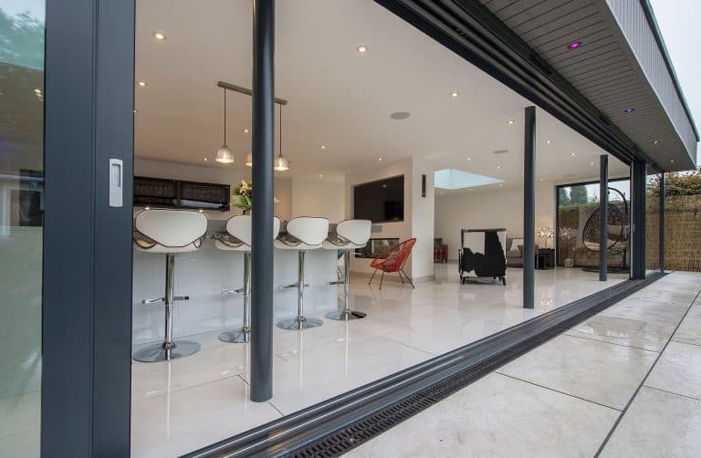 Modern open-plan kitchen and living area with white bar stools, pendant lights, tiled floor, and bespoke glass solutions like sliding doors, viewed from outside on a patio.