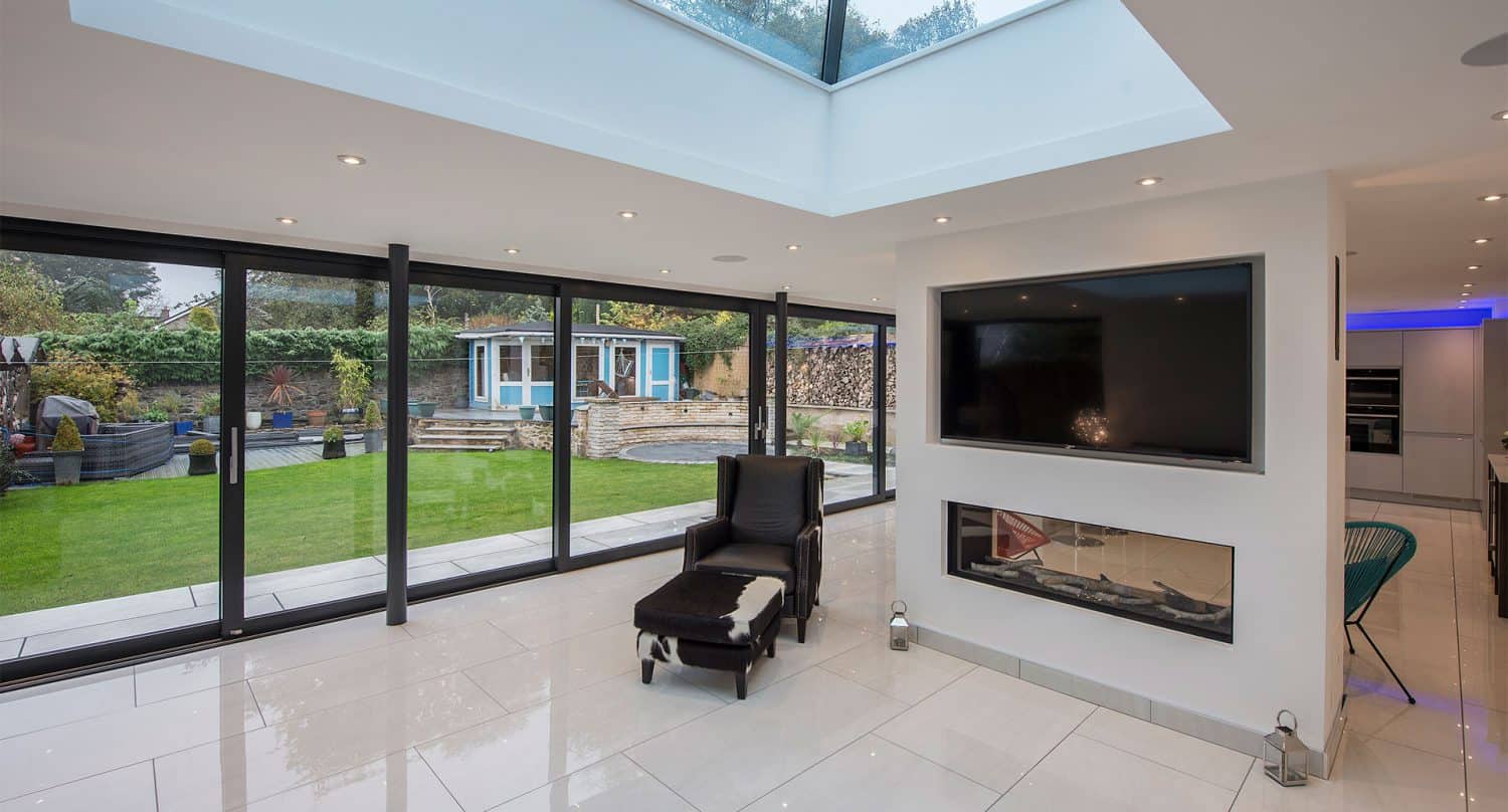 Modern living room with large bifold doors overlooking a green backyard, a skylight, a wall-mounted TV above a fireplace, and a black lounge chair with ottoman.