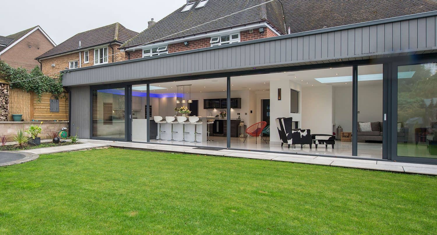 Modern house with expansive bifold doors opening onto a green lawn, revealing a spacious open-plan kitchen and living area with bar stools, armchairs, and contemporary decor inside.