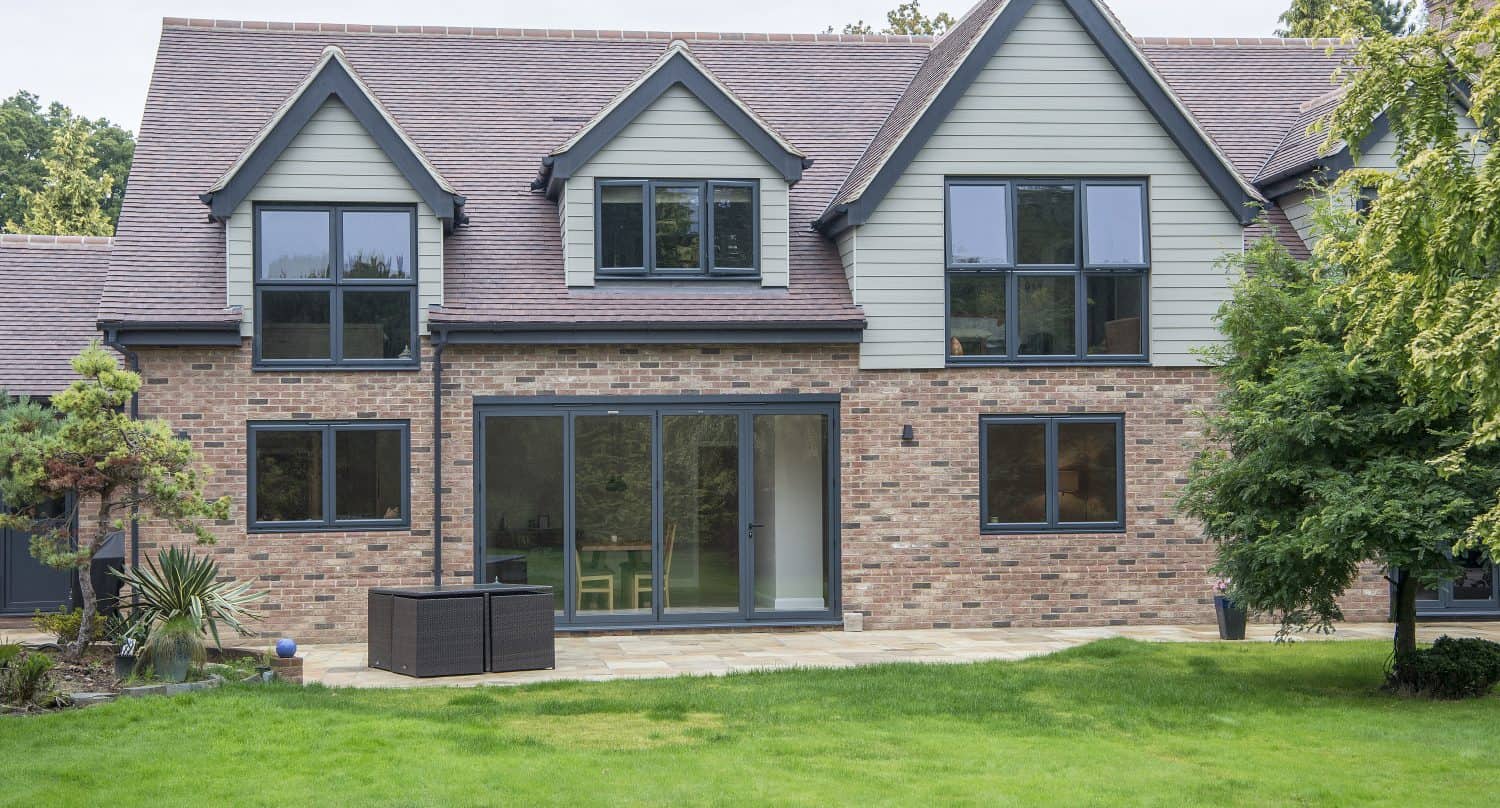 Modern two-story house with brick and light siding, large black-framed windows, and bifold doors opening to a spacious green backyard with a small patio and various plants.