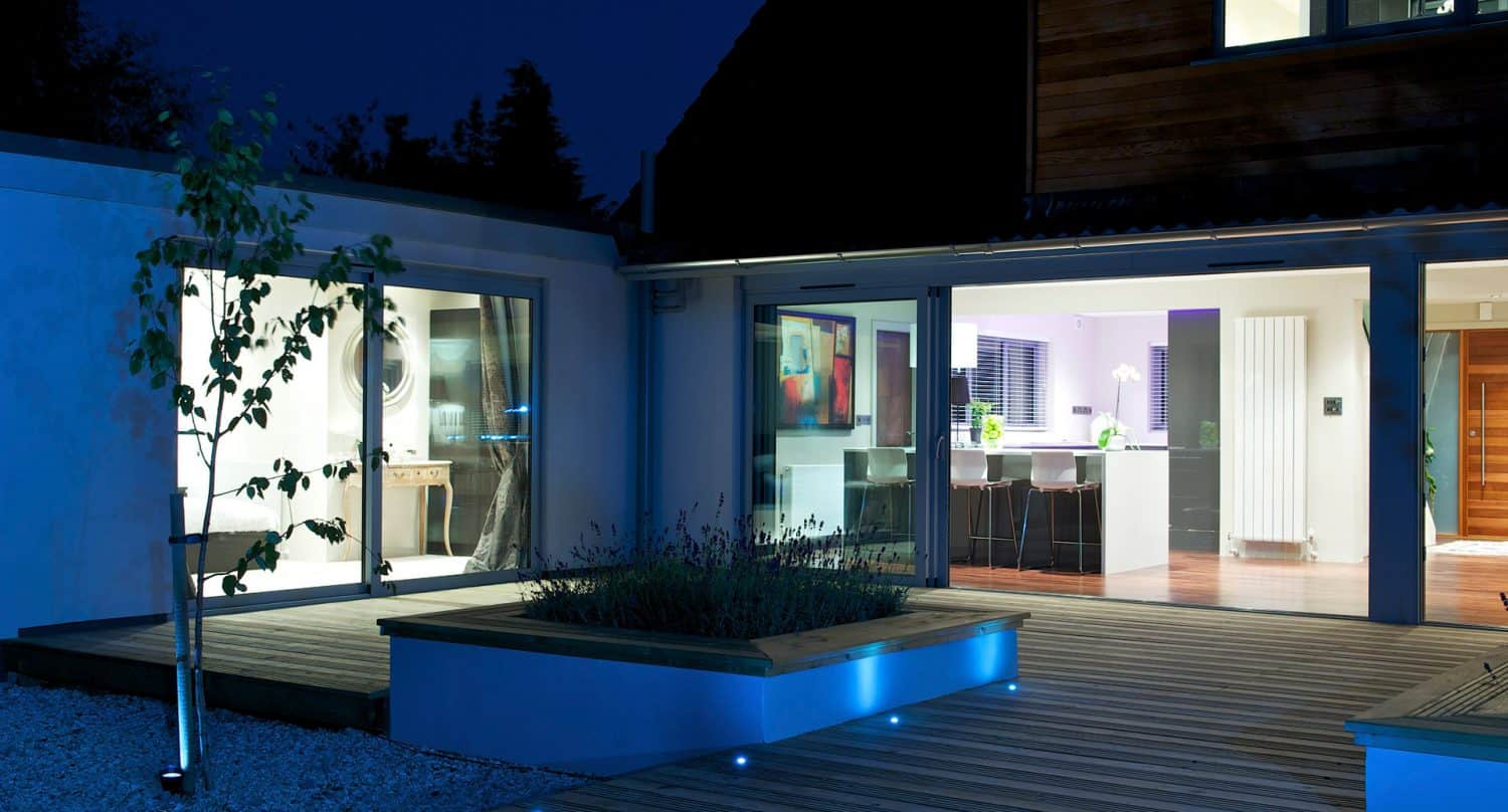 A modern house at night with large glass doors opening to a wooden deck, warmly lit interior showing a kitchen and dining area, and outdoor planters with blue accent lighting.