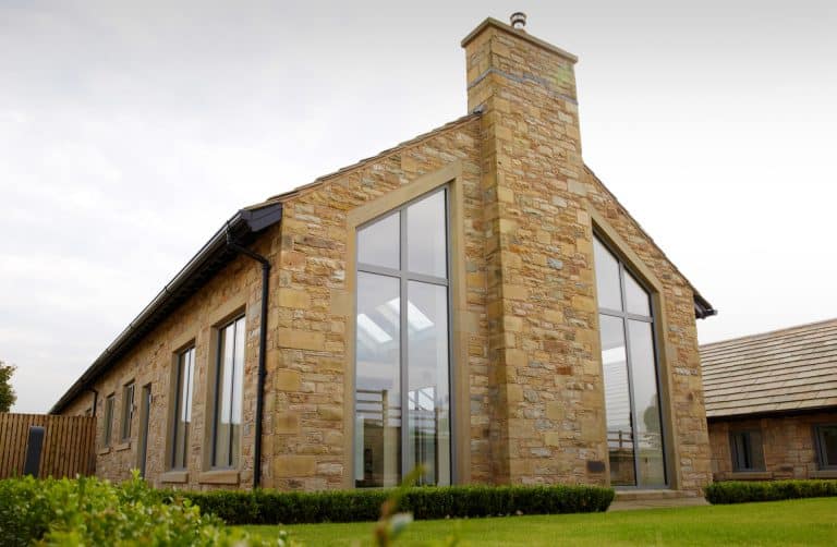 A modern house with stone exterior walls, large floor-to-ceiling windows, a chimney, and a well-maintained lawn in the foreground.