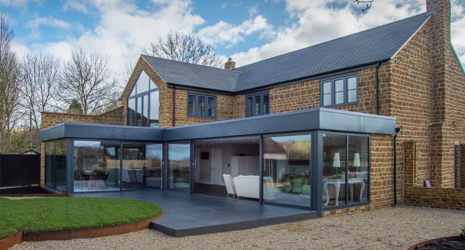 Modern extension with large glass windows and patio attached to a traditional stone house, surrounded by gravel and grass, with trees and a partly cloudy sky in the background.