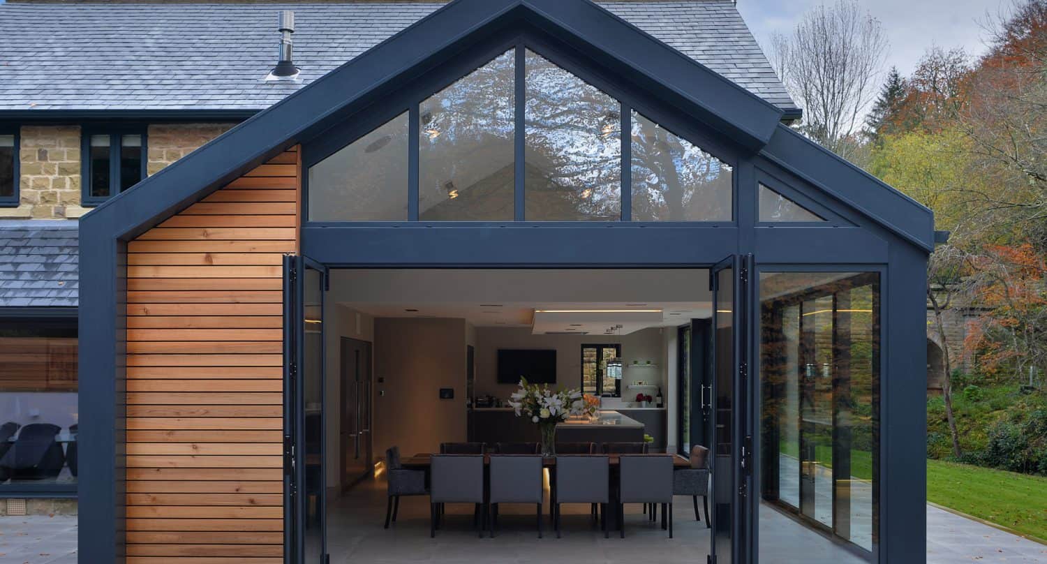 Modern house extension with large glass panels, folding glass doors, and wooden accent. The interior features a dining table with chairs and a kitchen area, opening to a patio in a green, wooded setting.