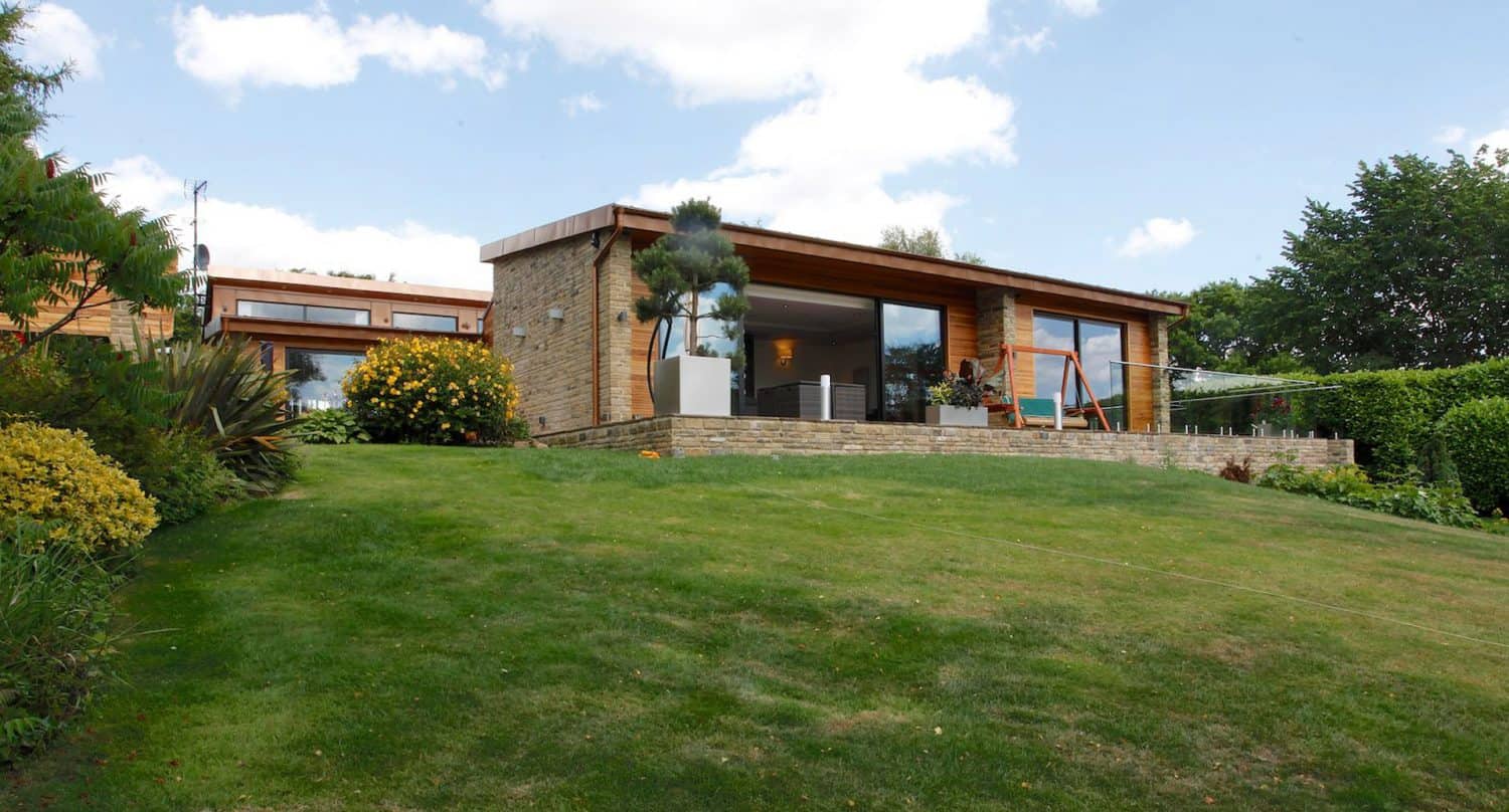 A modern, single-story house with large glass windows and wood accents sits on a grassy lawn, surrounded by green shrubs and trees under a partly cloudy sky.