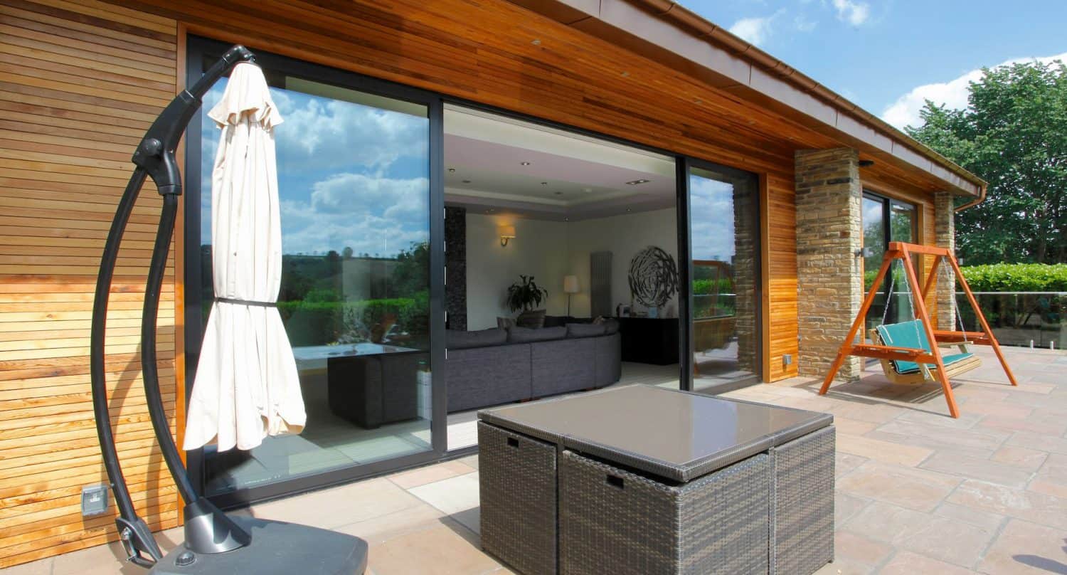 Modern patio with wicker furniture, a closed umbrella, and a wooden swing set. Large glass sliding doors open to a living room with a gray sofa. The house exterior is wood-paneled with a view of greenery outside.