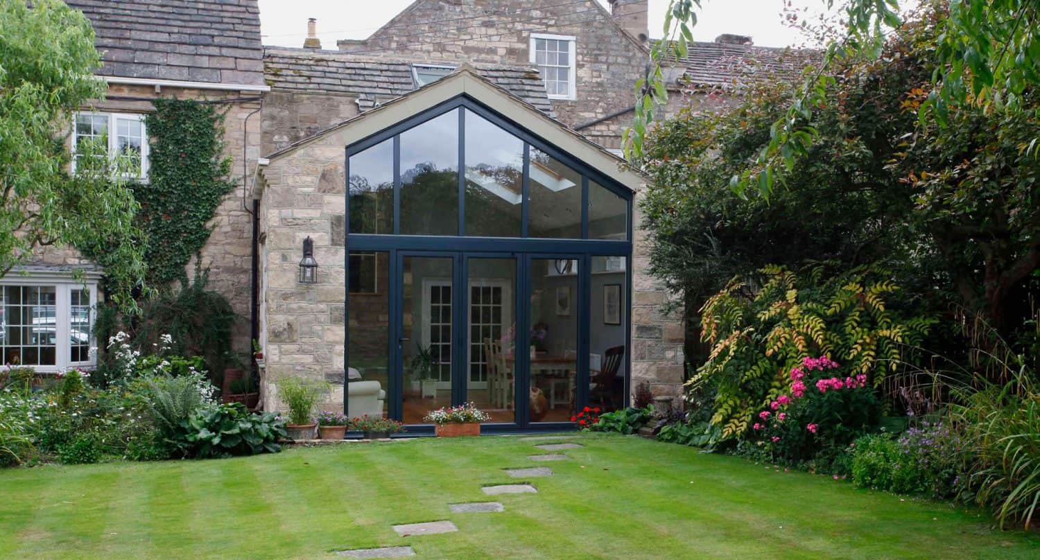 A stone house features a modern glass extension with a high, triangular roof. The garden has a green lawn, stepping stones, colorful flowers, and lush plants surrounding the house.