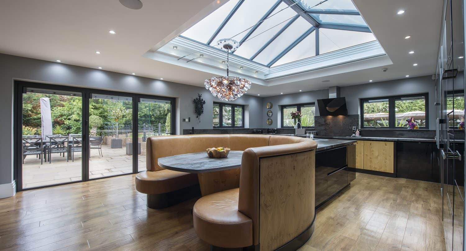 Modern kitchen with bespoke glass solutions, including a large skylight, wooden floors, black and wood cabinetry, a curved tan leather booth around a round table, and glass doors opening to a patio with outdoor seating and greenery.