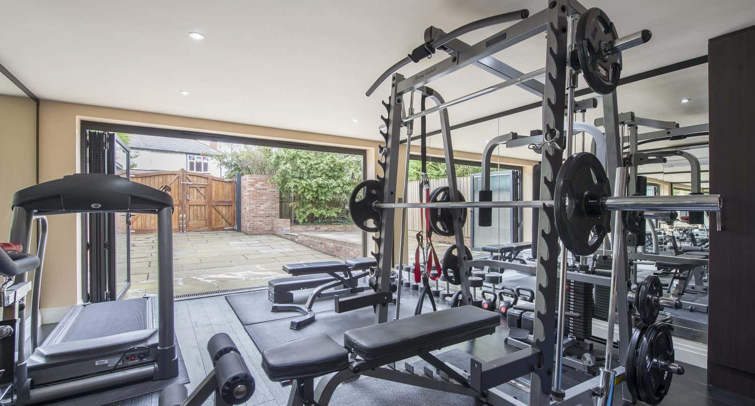 A modern home gym with weightlifting equipment, bench, treadmill, and large windows with bifold doors opening onto a patio and garden. The space is bright, well-organized, and perfect for energizing workouts.