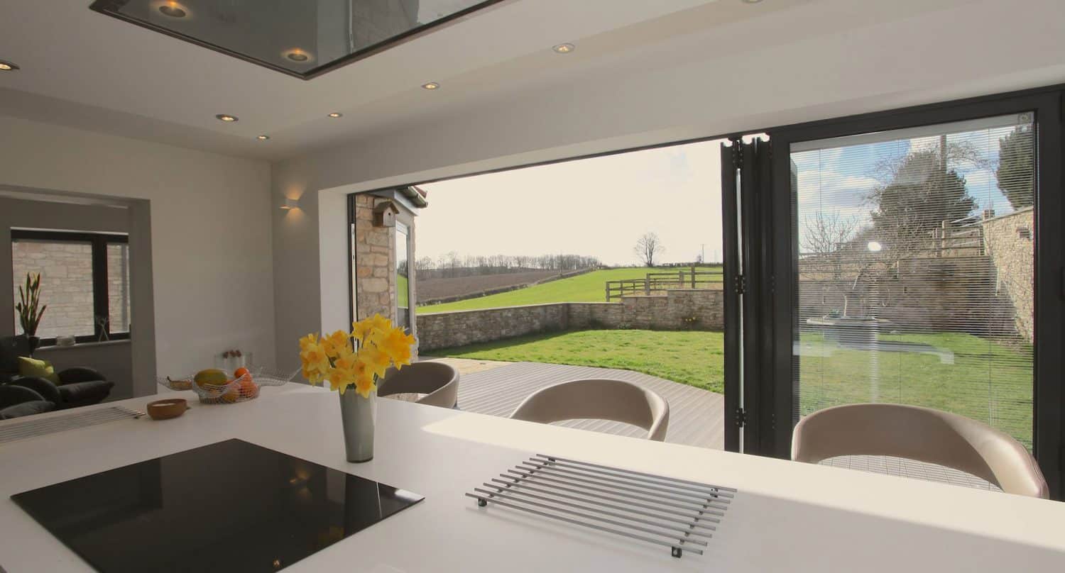 Modern kitchen with a white countertop, induction stove, and a vase of yellow flowers, opening to a sunny patio and green lawn through large glass folding doors.