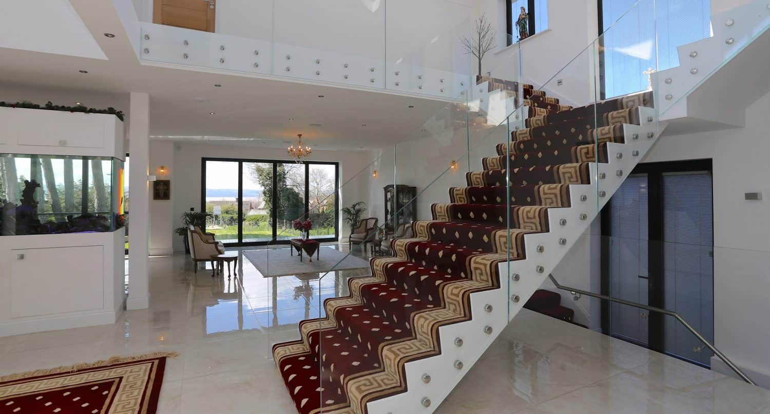 Modern, spacious living area with large windows, glossy tiled floors, and a red carpeted staircase with glass railings. The room features a chandelier, elegant chairs, and views of greenery outside.