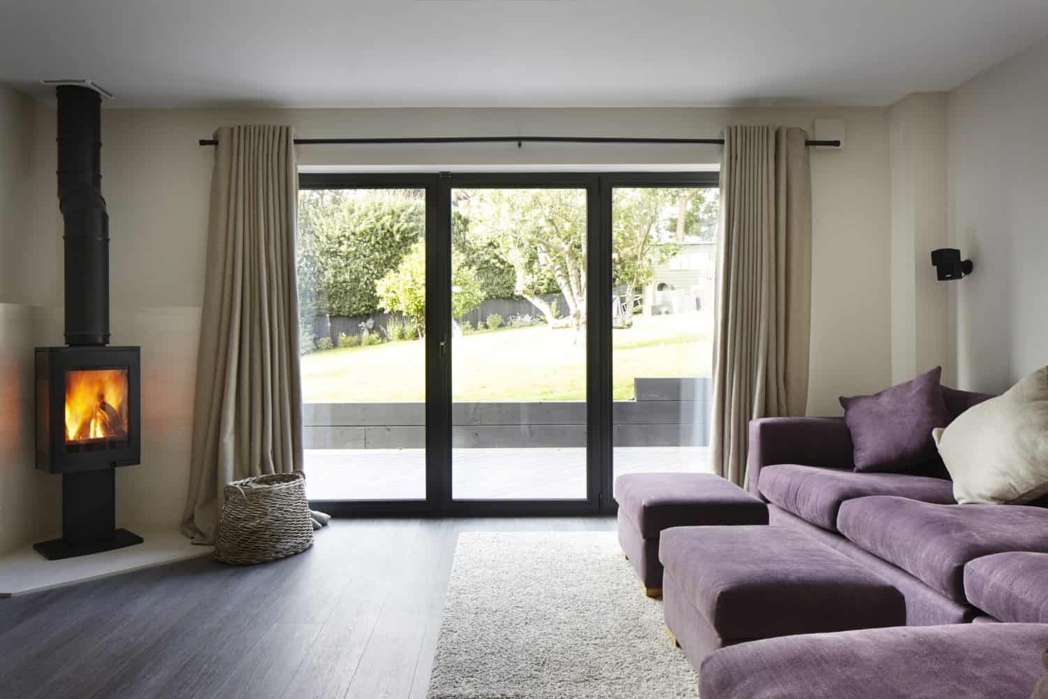 Modern living room with a purple sectional sofa, wood-burning stove, woven basket, light rug, and large bifold doors with beige curtains offering a beautiful view of the sunny garden outside.