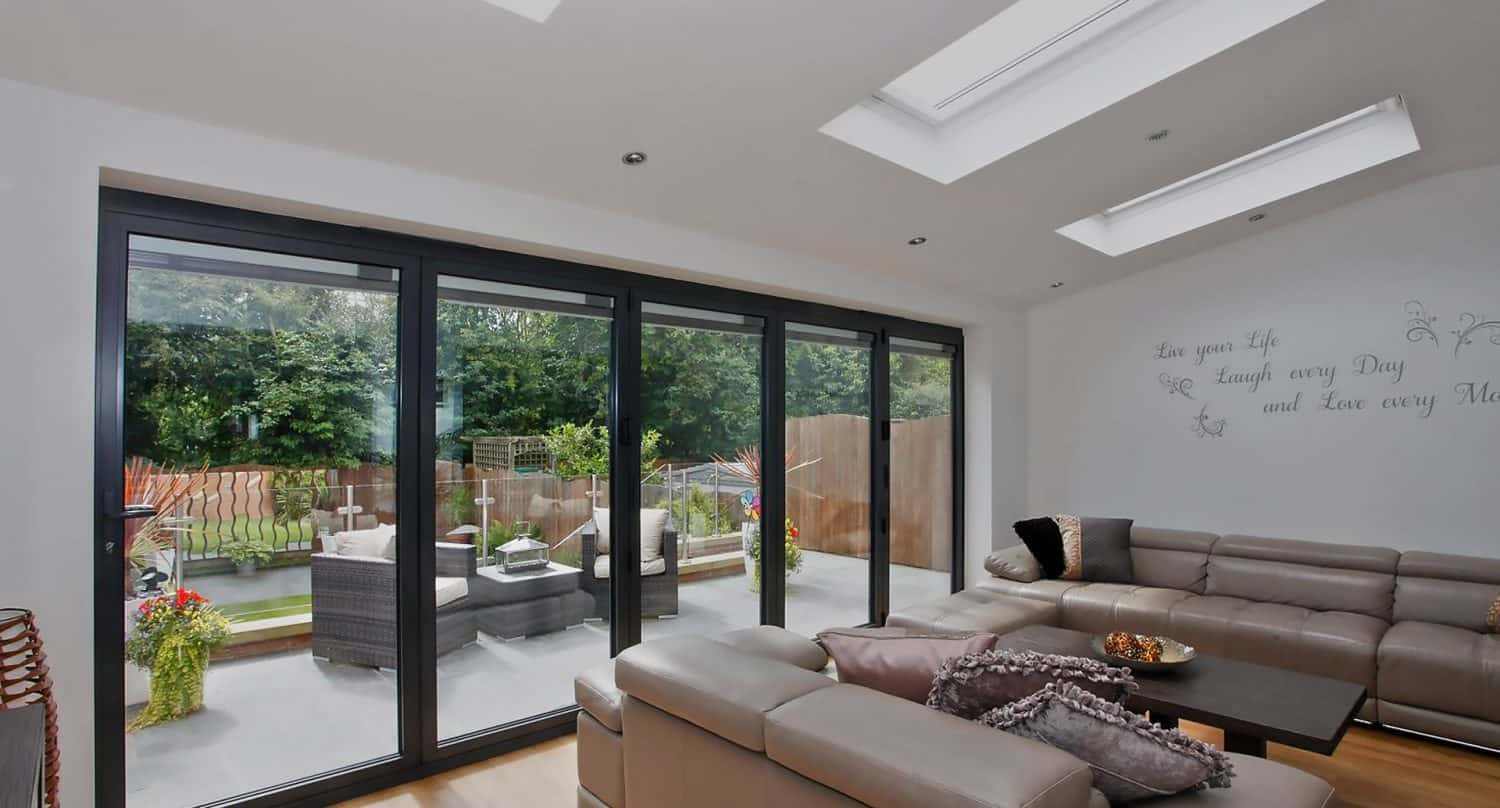 Modern living room with large glass sliding doors opening to a patio and garden. The room has beige leather sofas, decorative pillows, and wall art. The patio features outdoor seating and green trees in the background.