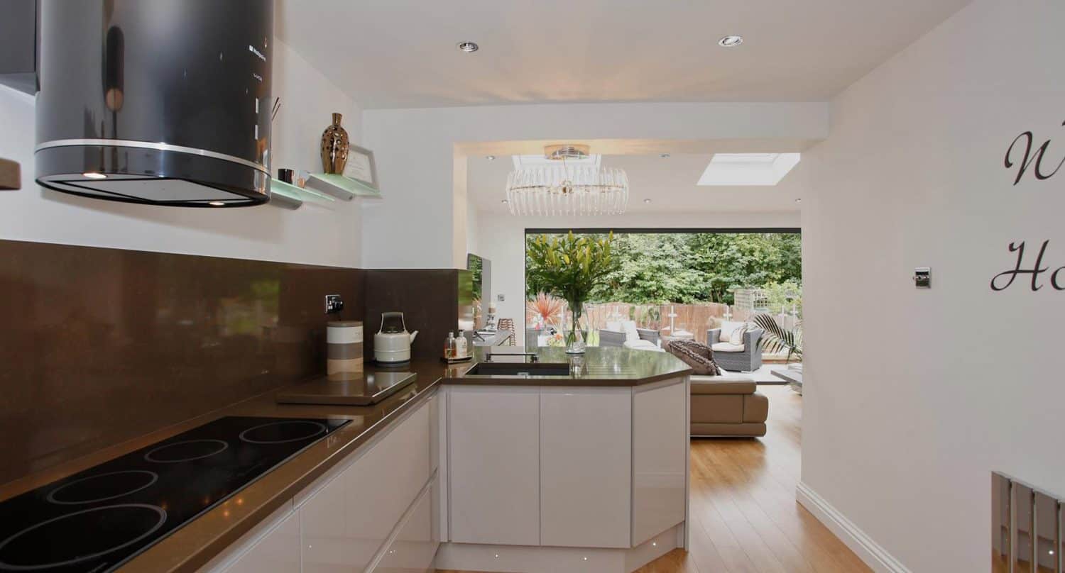 Modern kitchen with glossy cabinets, black stovetop, and brown backsplash, opening into a bright living room with large windows, wooden floors, a chandelier, and a view of a green garden outside.