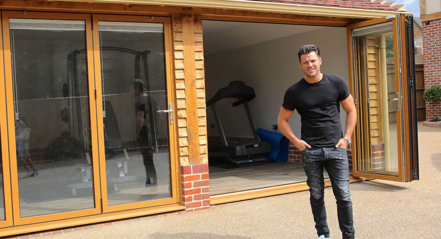 A man in a black t-shirt and jeans stands outside a modern home gym with large glass doors, with fitness equipment like a treadmill visible inside.