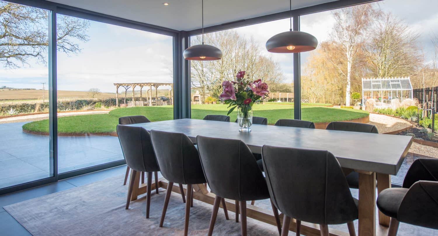 A modern dining room with a large wooden table, eight dark chairs, two pendant lights, and a vase of flowers. Floor-to-ceiling windows reveal a garden and expansive green lawn outside.