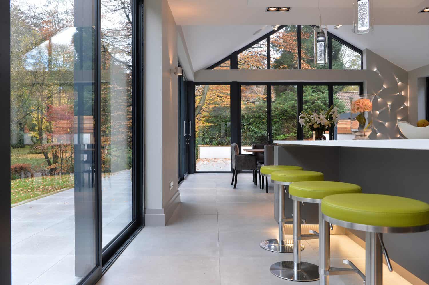 Modern kitchen with large glass windows and bespoke bifold doors overlooking a garden with autumn trees. Green bar stools line a white island, and natural light fills the space. A dining table is visible in the background.