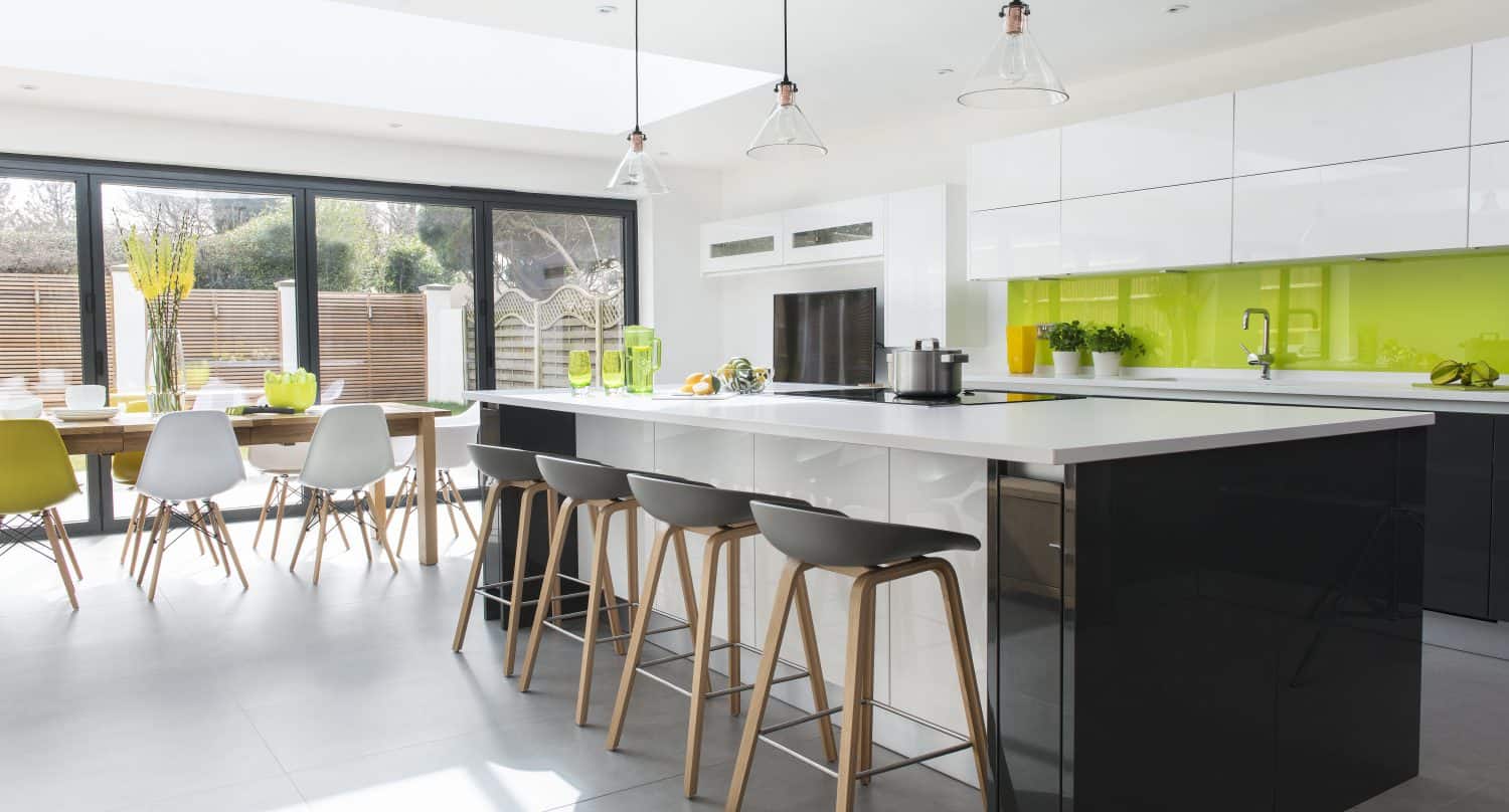 Modern kitchen with a large island, bar stools, white cabinets, black lower drawers, and a bright green glass backsplash. Sunlit dining area with wooden table and chairs is visible through expansive bifold doors.