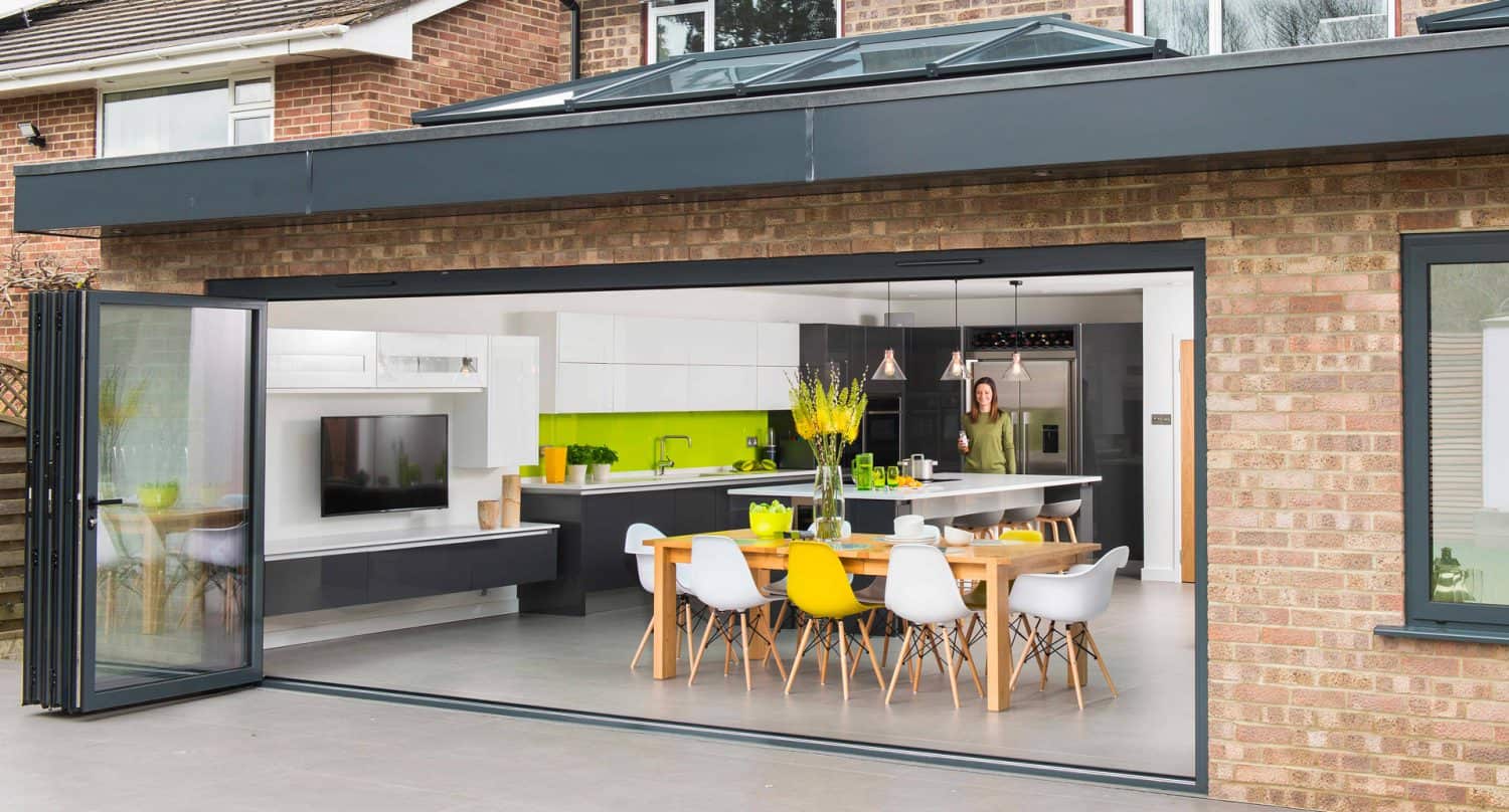 A modern open-plan kitchen and dining area with large sliding glass doors, a wooden dining table with white chairs, green and yellow accents, and a person standing near the kitchen in the background.
