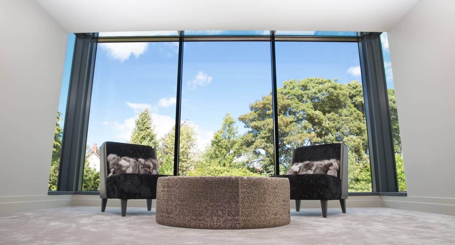 Two modern armchairs with patterned cushions and a round ottoman sit in a bright room with large floor-to-ceiling windows and bifold doors overlooking green trees and a blue sky with clouds.