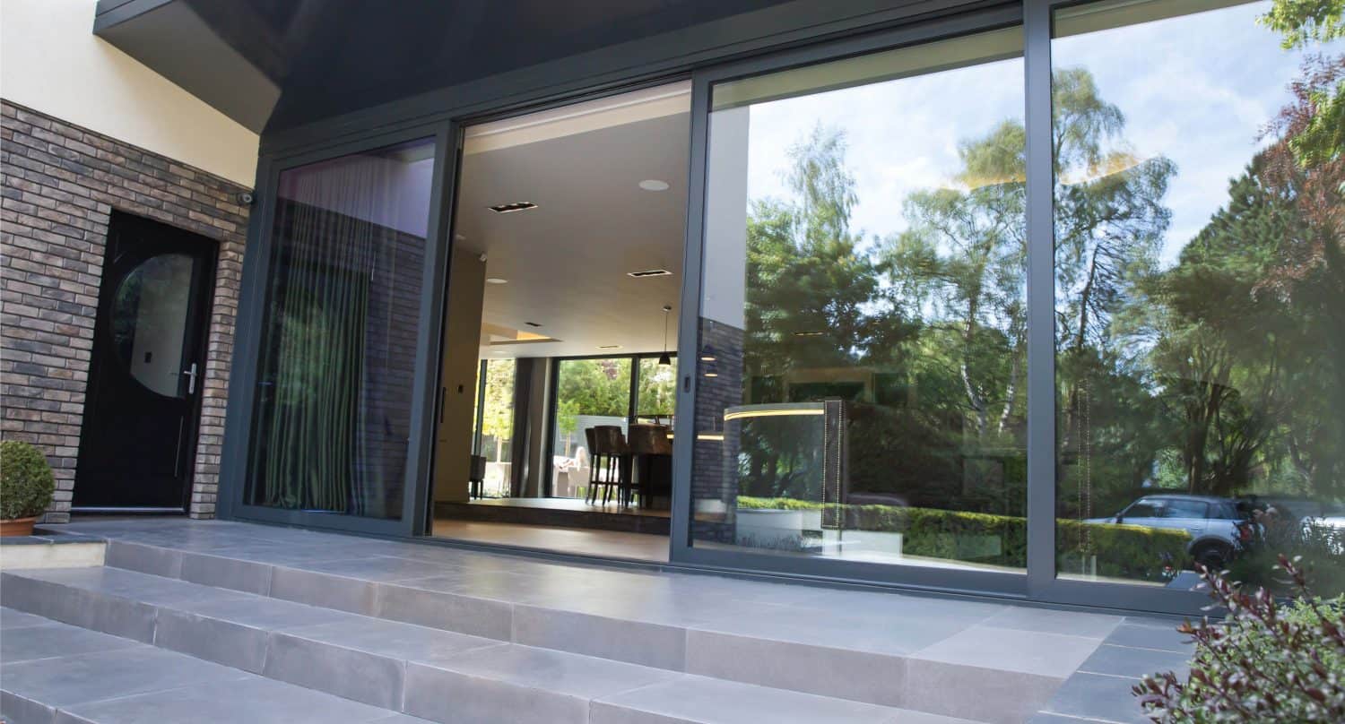 Modern house exterior with large sliding doors opening to a patio with gray tiled steps. The interior dining area is visible through the doors, and greenery is reflected in the glass.