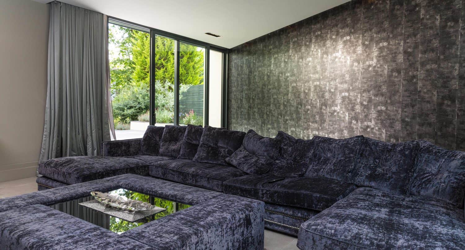A modern living room with a large, dark gray velvet sectional sofa, a matching ottoman with a glass tray, textured metallic accent wall, floor-to-ceiling curtains, and sliding doors opening to greenery.